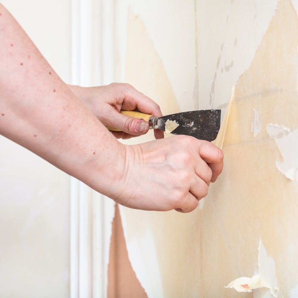 Mur en plâtre partiellement dégagé, avec une bande de papier peint soulevée et des outils de rénovation au sol.