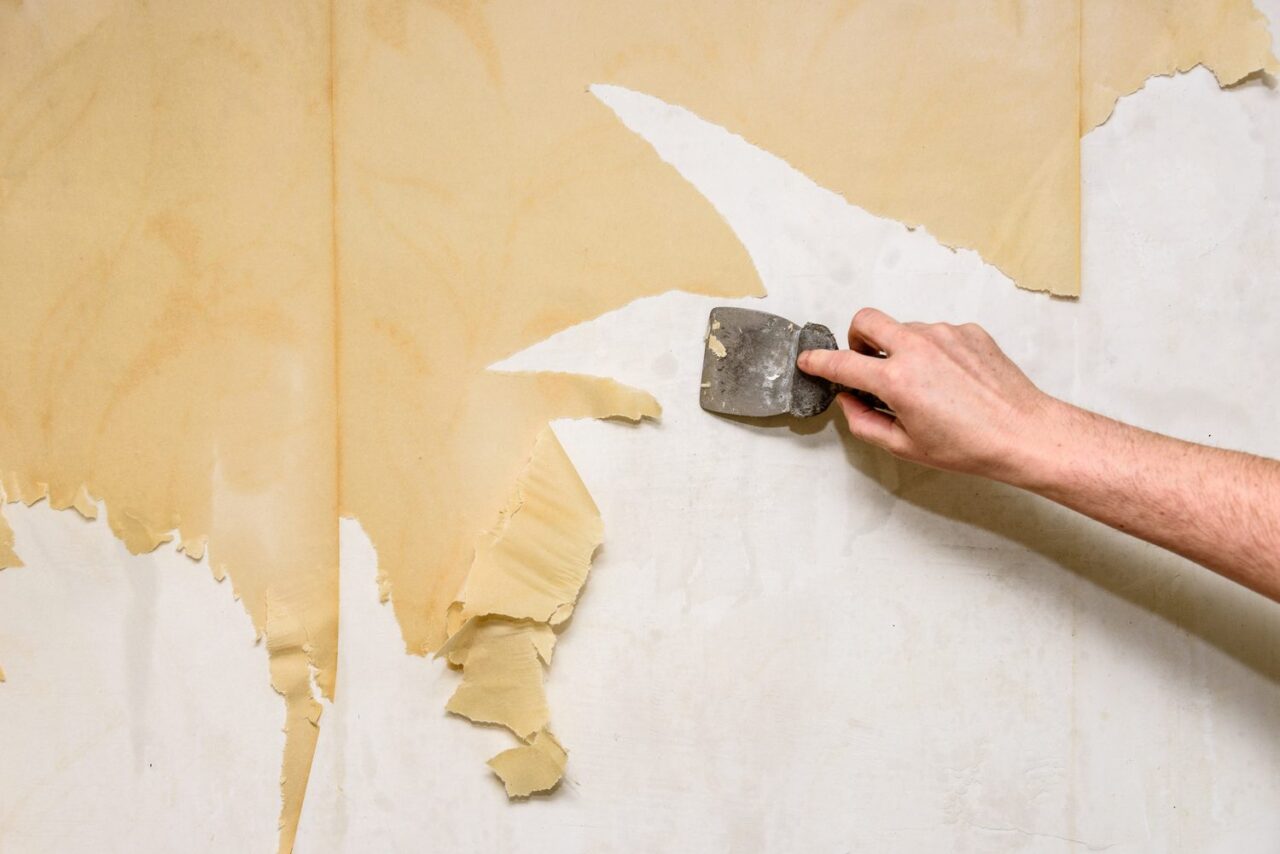 Personne retirant délicatement du papier peint d’un mur en plâtre à l’aide d’une spatule.